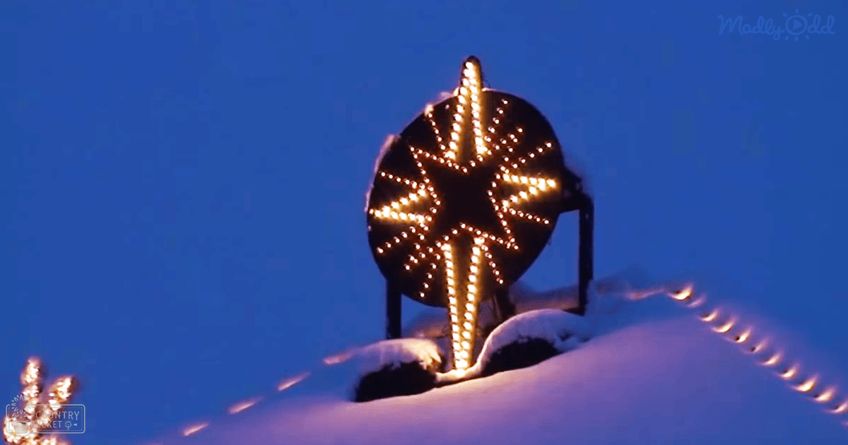 A symmetrical snowflake-shaped light display with golden-yellow lights on a snowy outdoor building at dusk.