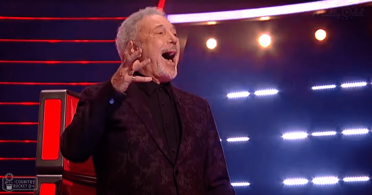 A middle-aged adult male wears a dark patterned blazer over a black shirt with buttons down the front, seated on a red chair against blue studio lighting and red accents.