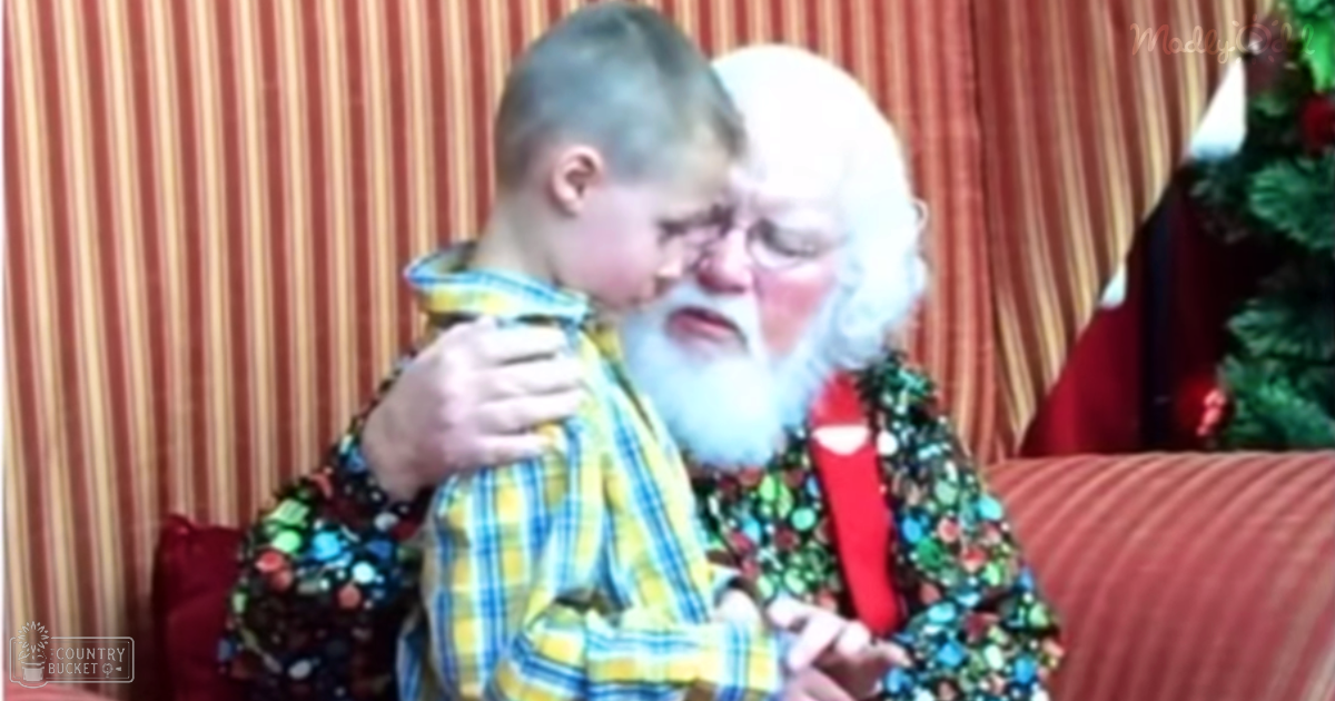 A toddler boy wearing a yellow and blue plaid shirt being hugged by an older adult dressed as Santa Claus in a home setting with a Christmas tree visible, indicating winter season.