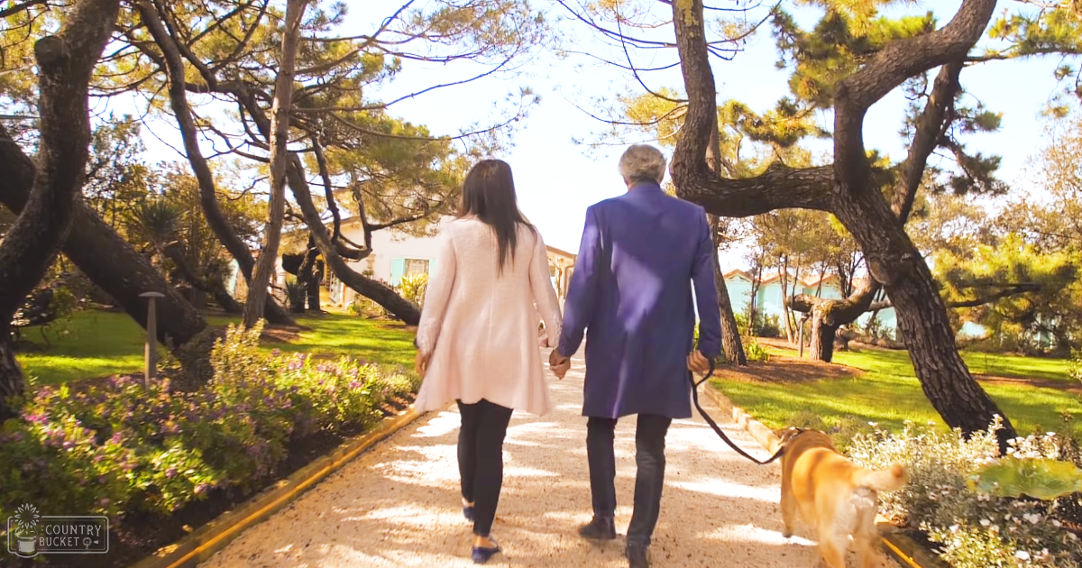 A couple walking their golden retriever on a garden path, holding hands under late afternoon light with green foliage and blue sky as backdrop.
