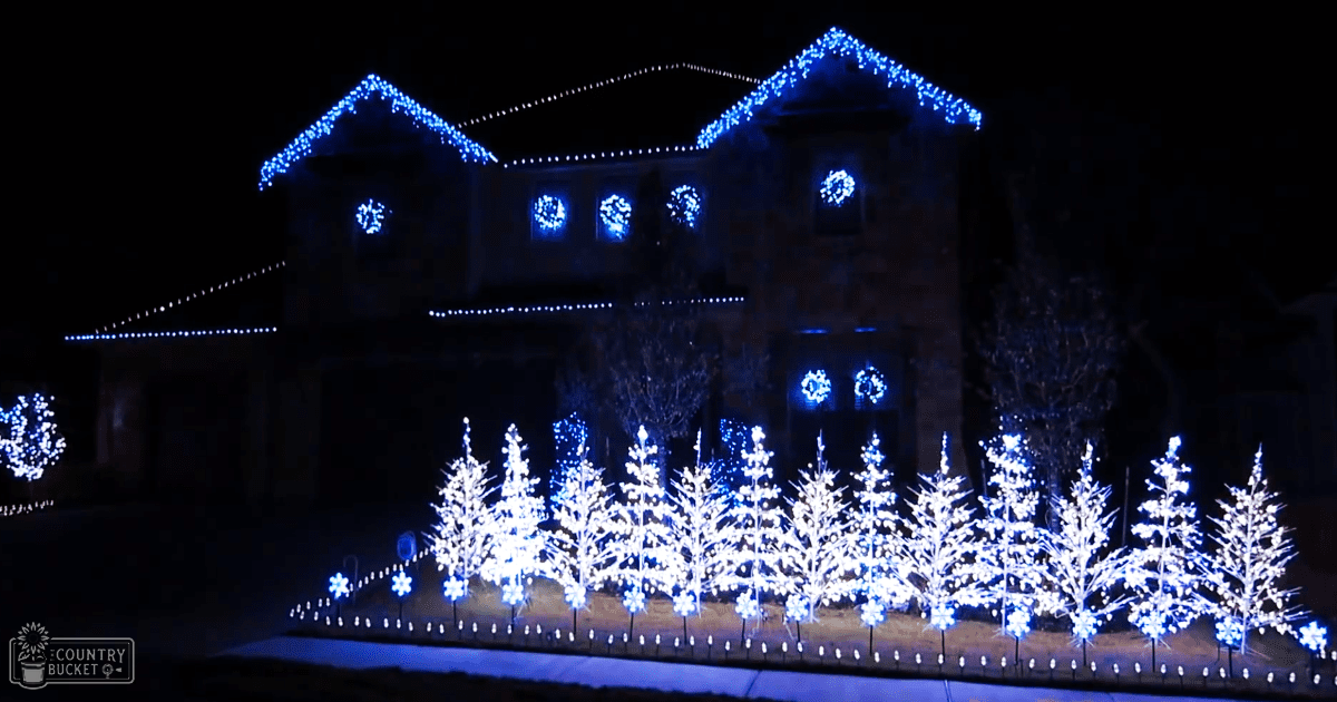 A large stone house adorned with numerous blue Christmas lights on its roof and windows, surrounded by small trees decorated with white snowflake-shaped lights at night in a winter urban residential setting.