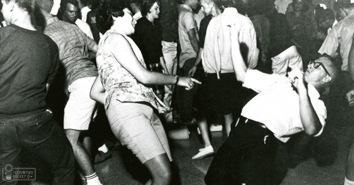 A crowded dance floor scene in an indoor setting, possibly a nightclub or social gathering from the mid-20th century with people wearing casual attire dancing and socializing under artificial evening lighting.