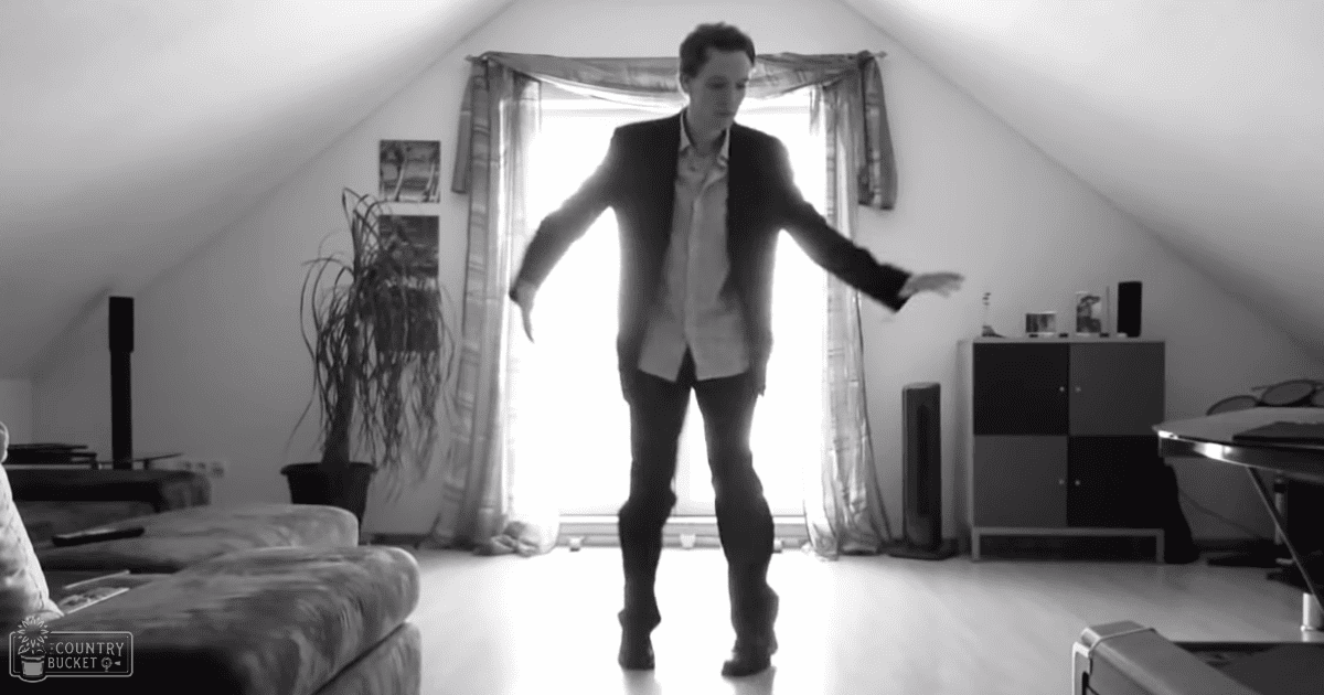 A young adult male wearing a tailored black blazer over a buttoned shirt with rolled-up sleeves, dark pants, and dark shoes stands amidst furniture including a patterned sofa, a potted plant, and an attic room's sloped ceiling under bright daylight.