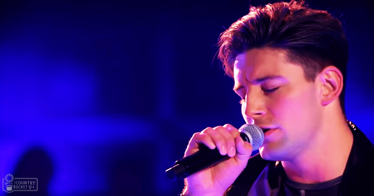 A young adult male in casual attire performs live music at an indoor concert venue with blue stage lighting.