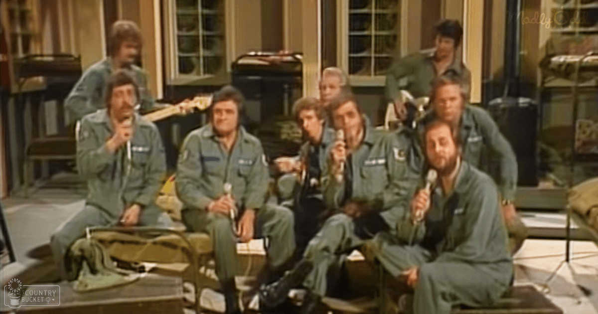 Performers in green military-style uniforms on an indoor stage with microphones and furniture, suggesting a comedy skit setup.