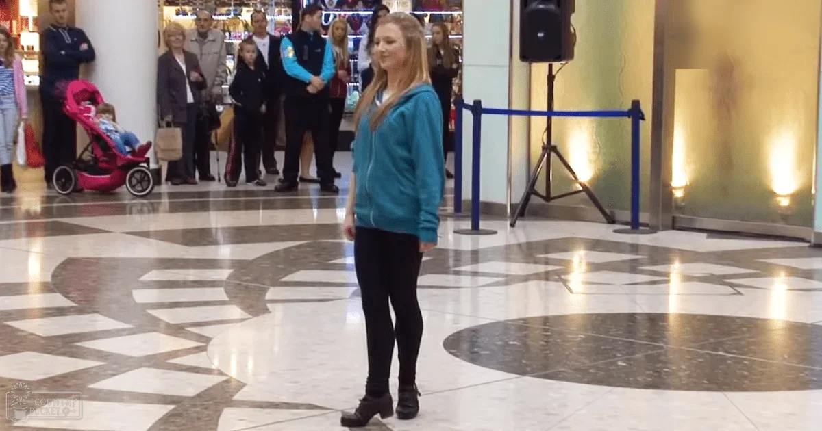 A young adult female wearing a teal zip-up hoodie and black leggings stands in the foreground of a busy shopping mall, surrounded by other shoppers including an older male with a stroller. A speaker on a tripod is visible to her right.