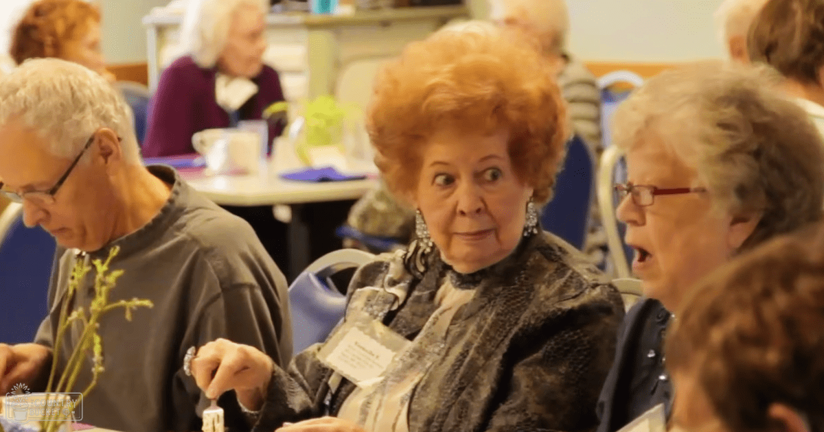 An older adult woman with red hair, wearing a patterned jacket in an indoor community center setting.