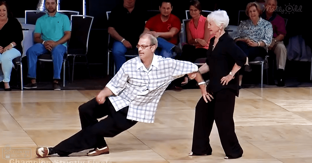 Middle-aged man dancing with an older adult woman in a dance studio or event space, surrounded by chairs and an audience watching.