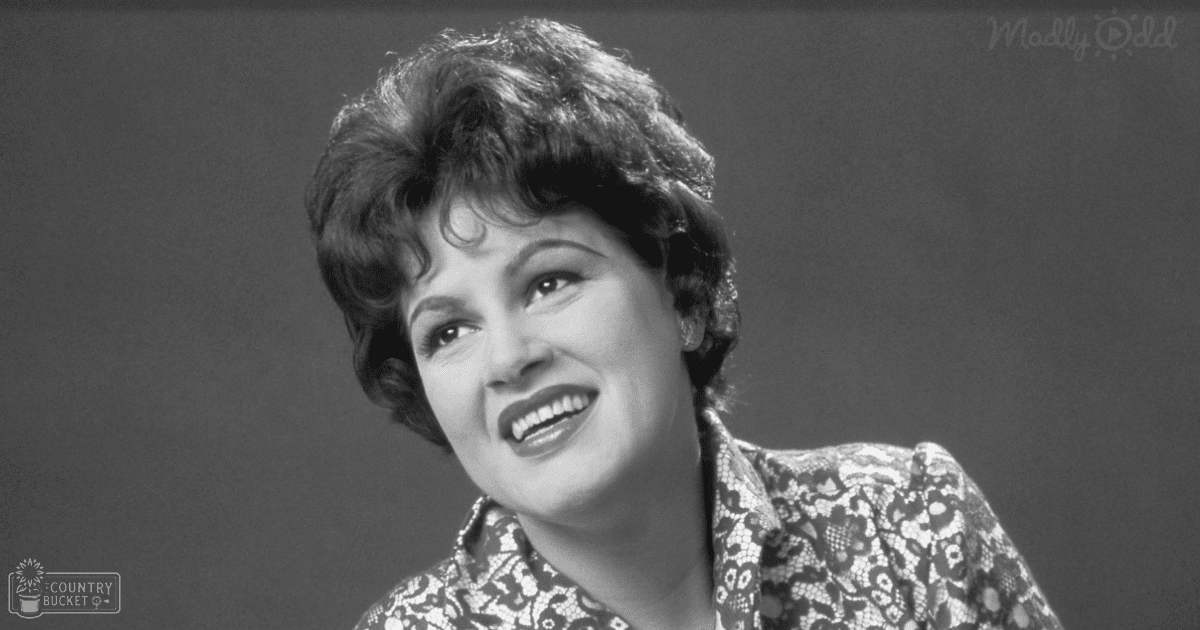 A middle-aged woman stands indoors in a monochrome patterned garment with short sleeves; the setting is a plain dark gray background, likely an indoor studio.