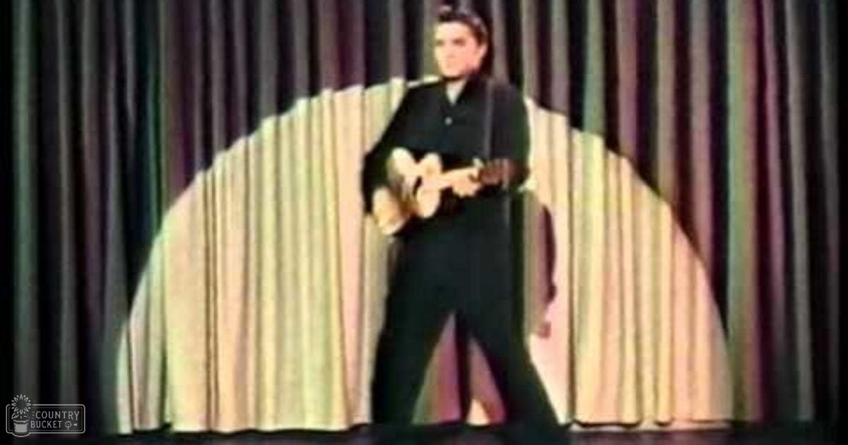A young adult male performer on stage, dressed in a dark suit and white shirt, holding a ukulele against the backdrop of a dark green stage curtain with vertical folds.