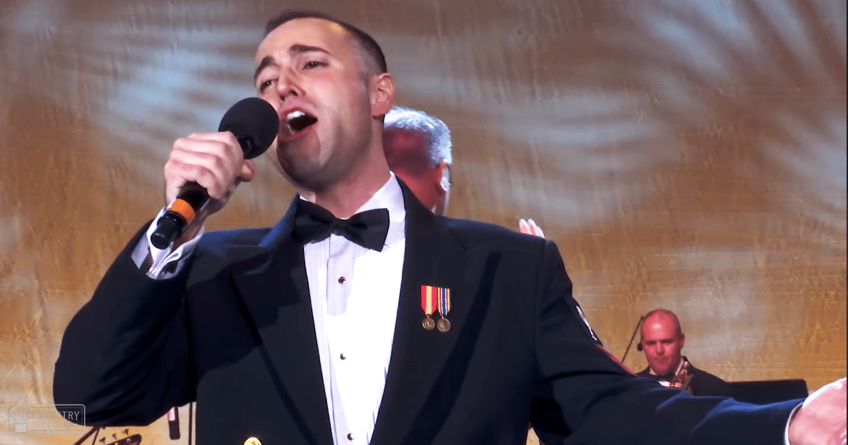 An adult male in a formal black tuxedo with medals, holding a microphone on stage at an indoor event.
