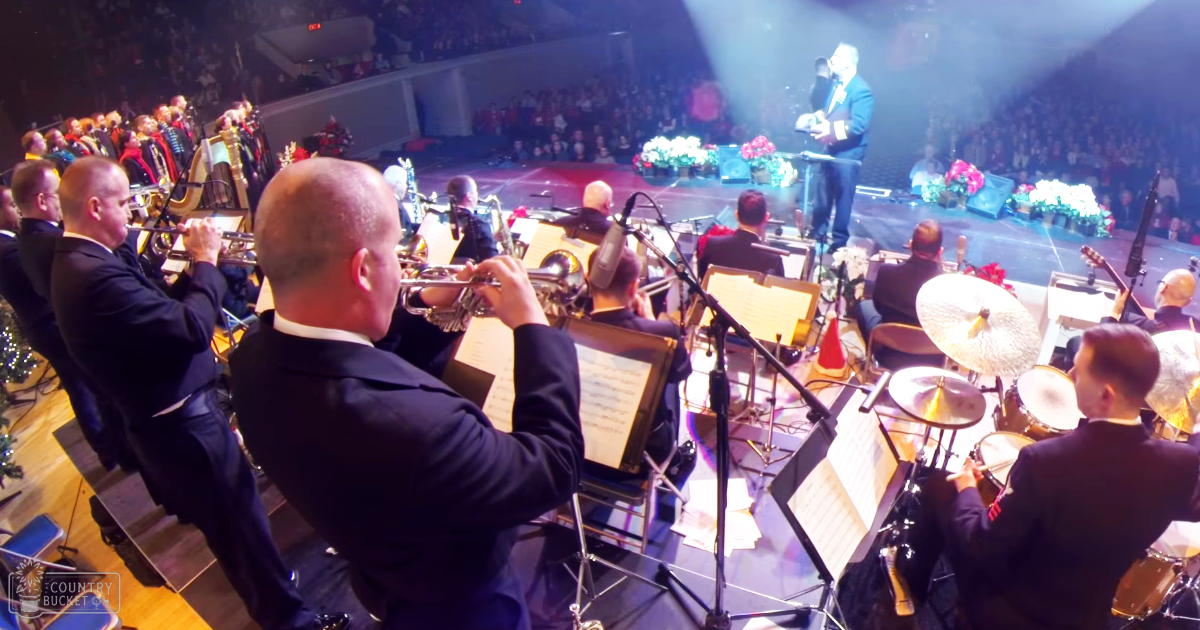 A live orchestra performance with adult male musicians wearing dark suits, white shirts, and black ties in an indoor concert hall setting.