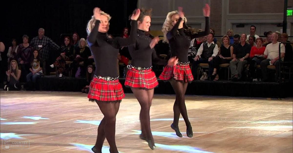 A group of young adult dancers performing Irish step dancing indoors with a seated audience.