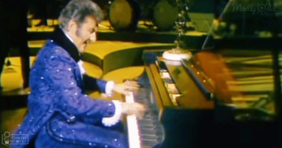 A male older adult performer wearing a blue ornate velvet coat and white gloves plays a grand piano on stage with dark curtains in an indoor theater setting.