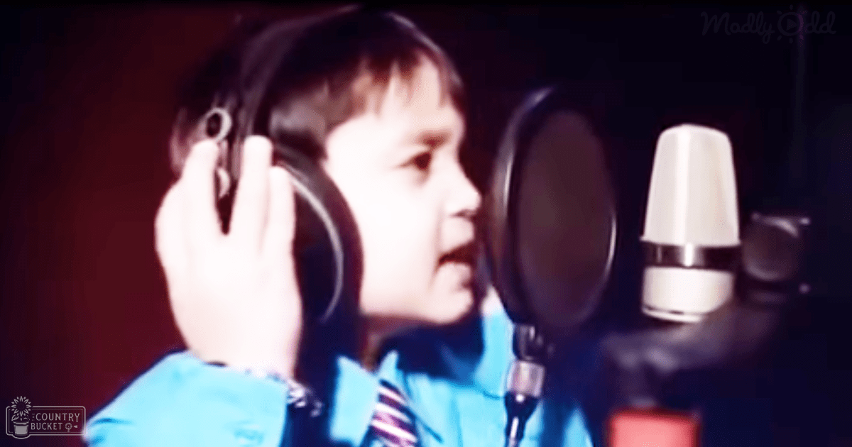 A young adult female wearing a light blue striped garment with dark hair styled neatly, standing in a recording studio with professional audio equipment.