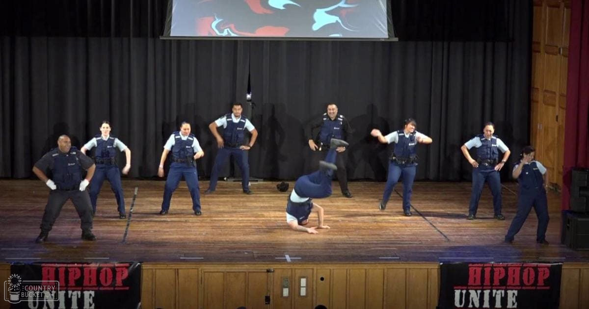 A group of young adults and middle-aged males participating in a hip-hop dance performance on an indoor stage, set against black curtains with 'HIPHOP UNITE' banners. The wooden floor and abstract art screen above the stage suggest a community or school hall.