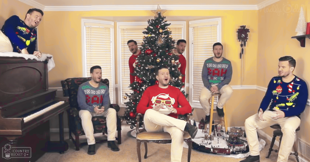 Group of young adults in festive indoor setting, likely during Christmas. Grand piano, decorated Christmas tree with lights and ornaments, cuckoo clock, small train set visible.