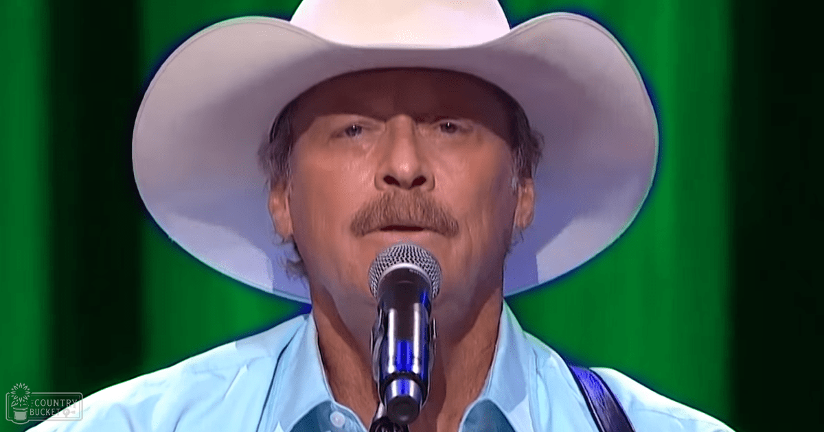 An older adult male in a performance setting, wearing a white cowboy hat and light blue button-up shirt, holding a microphone against a bright green stage backdrop.