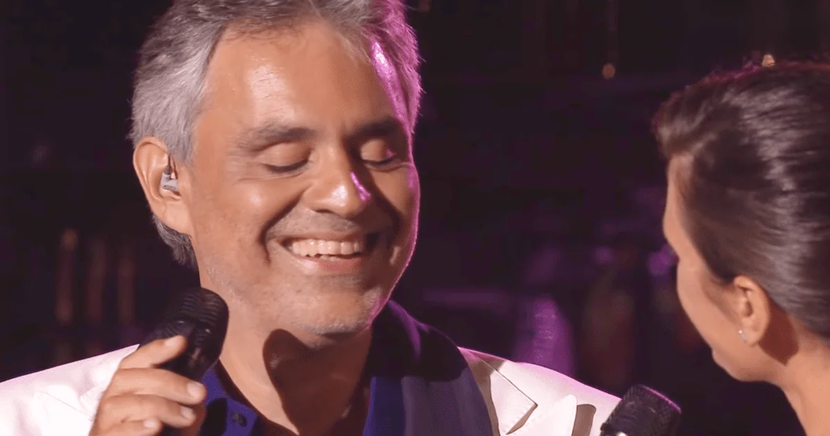 A middle-aged male performer in a white collared shirt with dark blue inner layer, holding a microphone on stage at what appears to be a concert or performance setting.