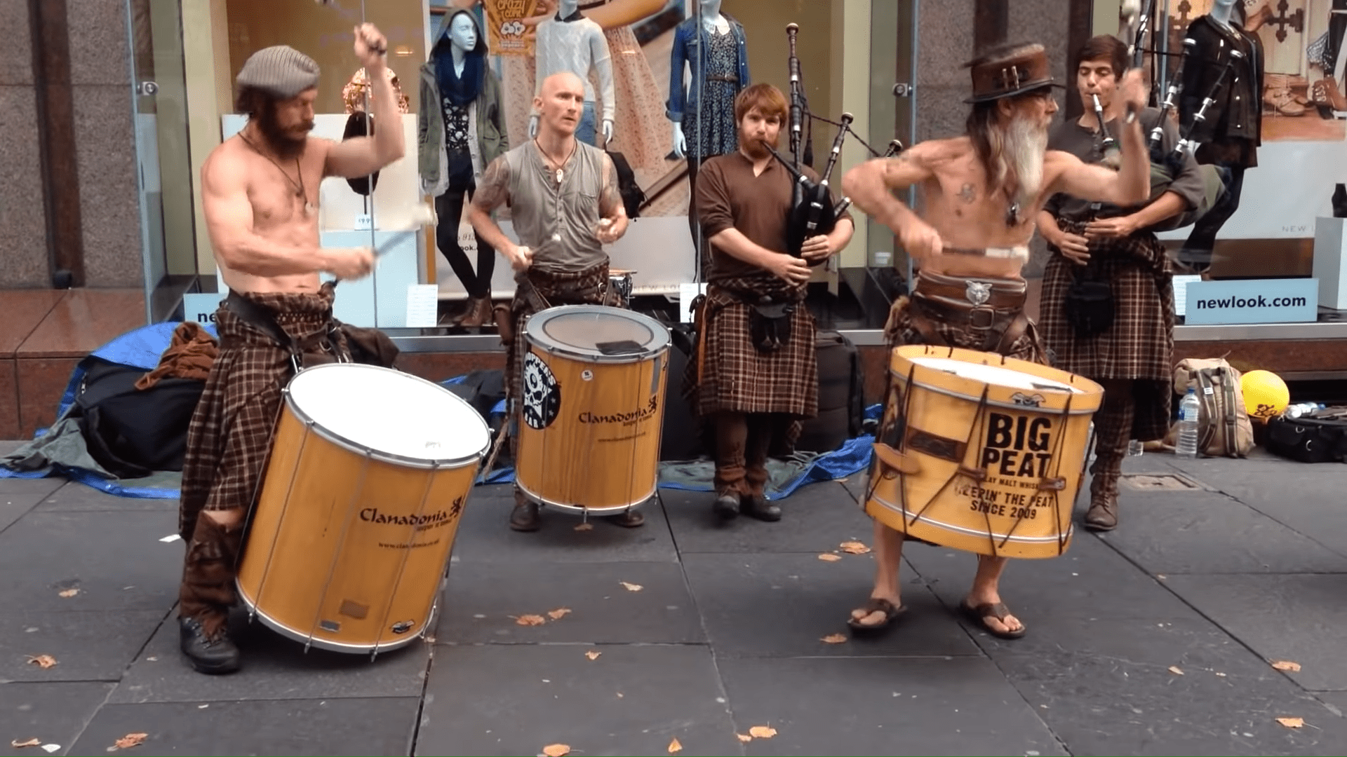 Street performance group in urban setting: adult males wearing kilts and playing drums, sign reading 'newlook.com' visible.