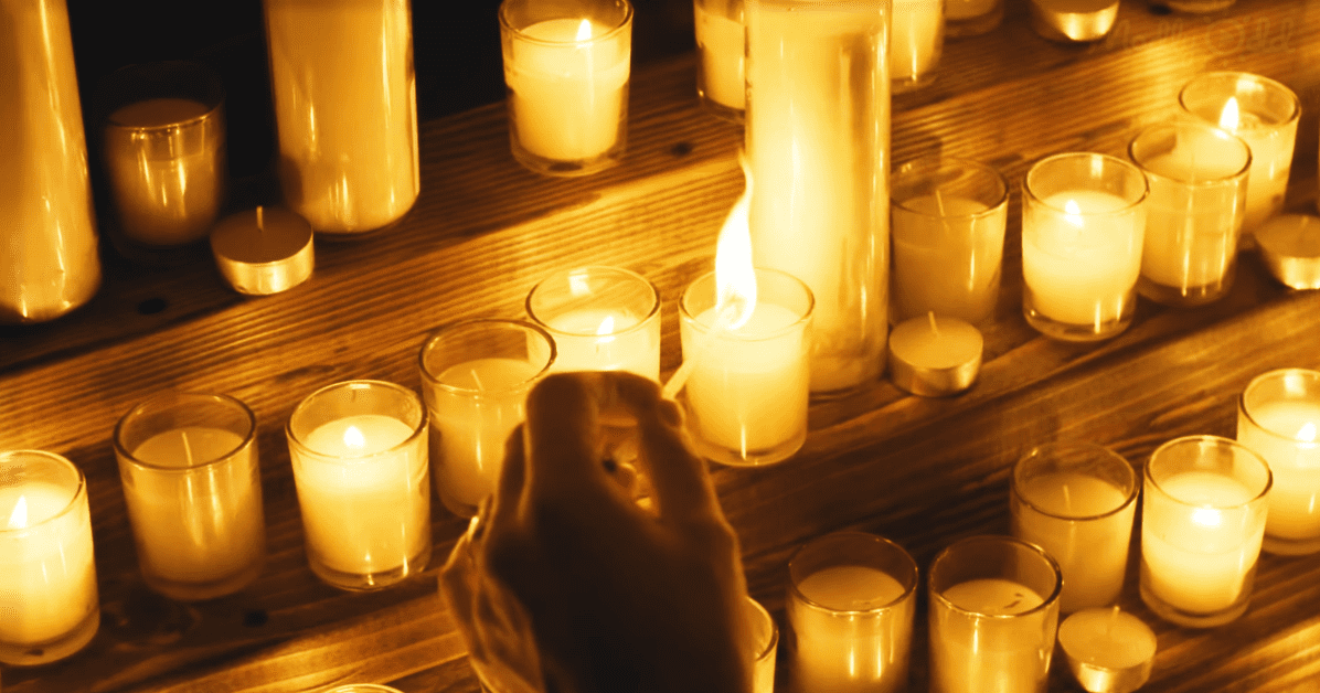 A hand lighting candles in an intimate memorial setting with warm yellow and orange candlelight casting a glow over the wooden surface.