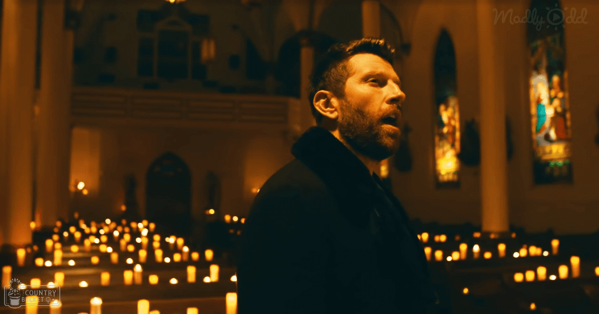 An adult male in a dark jacket with hood, inside an indoor church setting at night lit by candles and religious stained glass windows.
