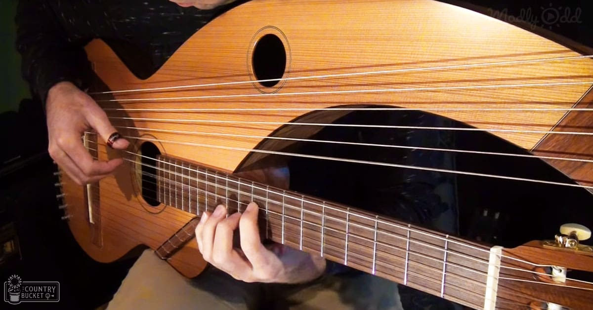 Adult male playing a wooden guitar indoors with artificial lighting, wearing a dark textured sweater or jacket.