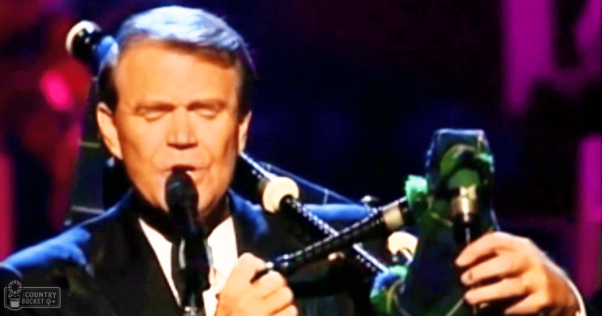 A middle-aged male performer wearing a dark suit jacket and white shirt with a tie plays green and black bagpipes in an indoor venue with dramatic purple lighting.