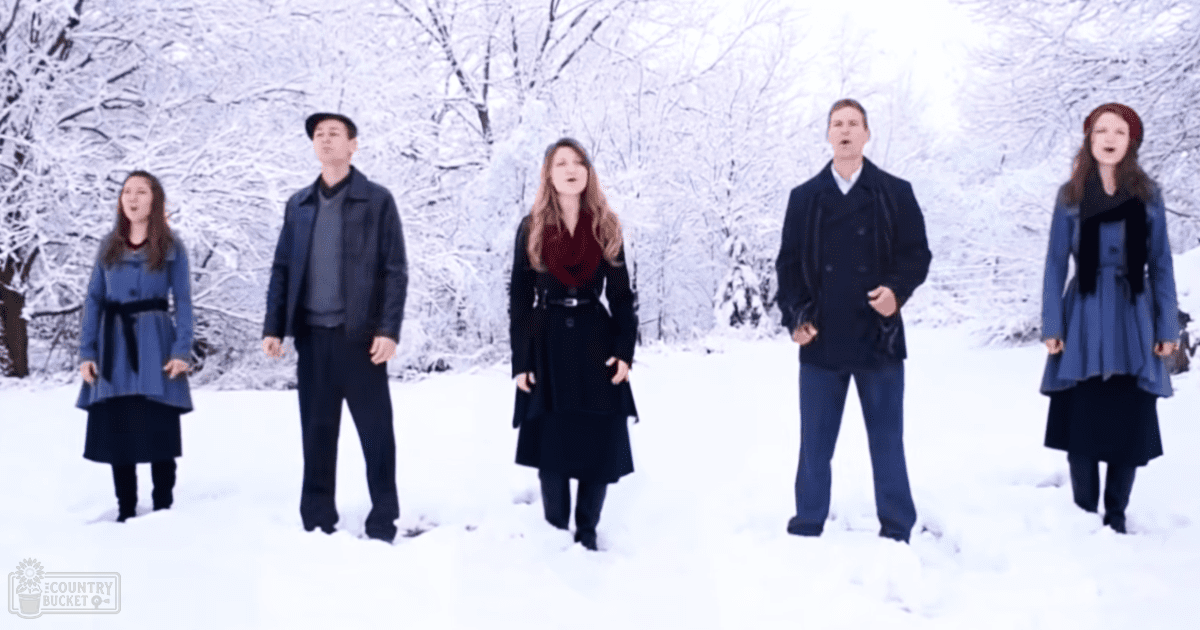Five adults singing outdoors in a snowy forest during winter, wearing dark blue coats and black boots, red scarf visible on one person.