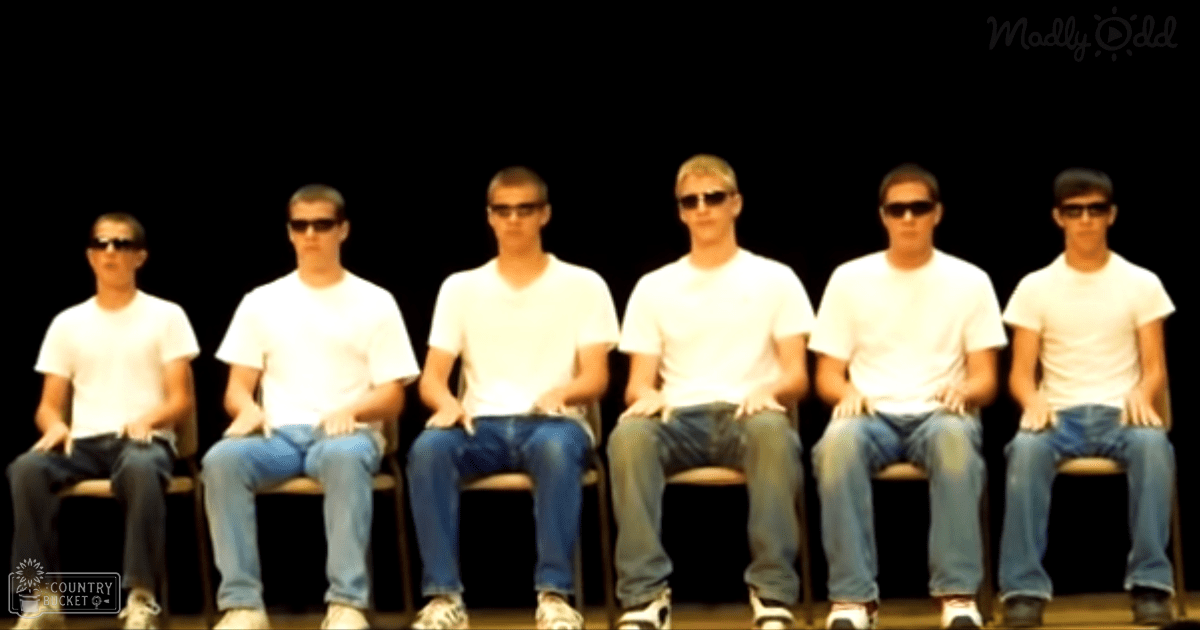 A group of young adult males in casual attire, wearing white t-shirts and dark/light blue jeans, seated on simple wooden chairs in an indoor stage setting with focused lighting.