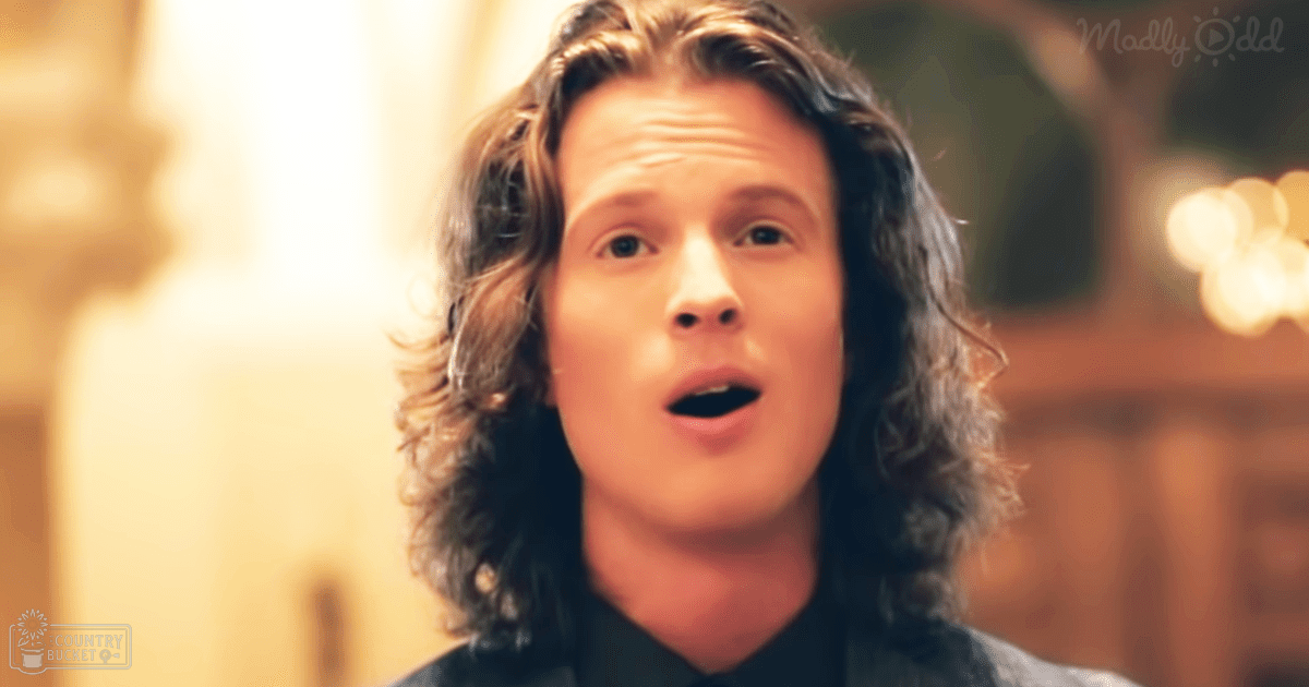 A young adult male wearing a dark-colored collared shirt indoors with warm lighting and ornate architectural details in the background.