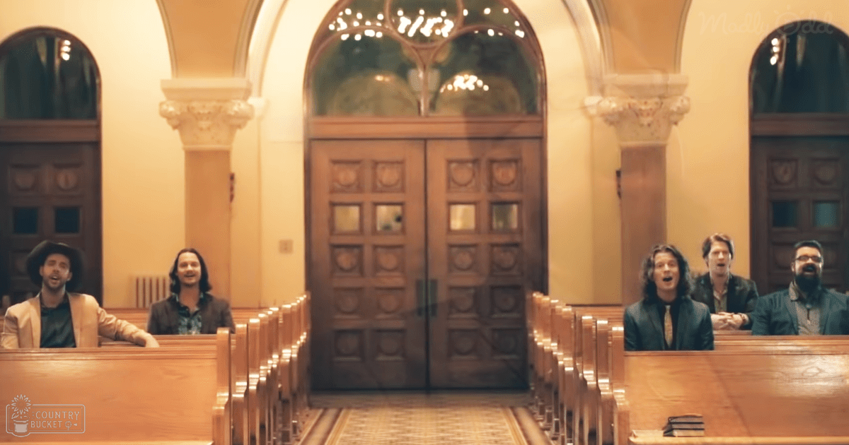 A group of adults singing in a church-like setting with arched glass doors and ornate columns.