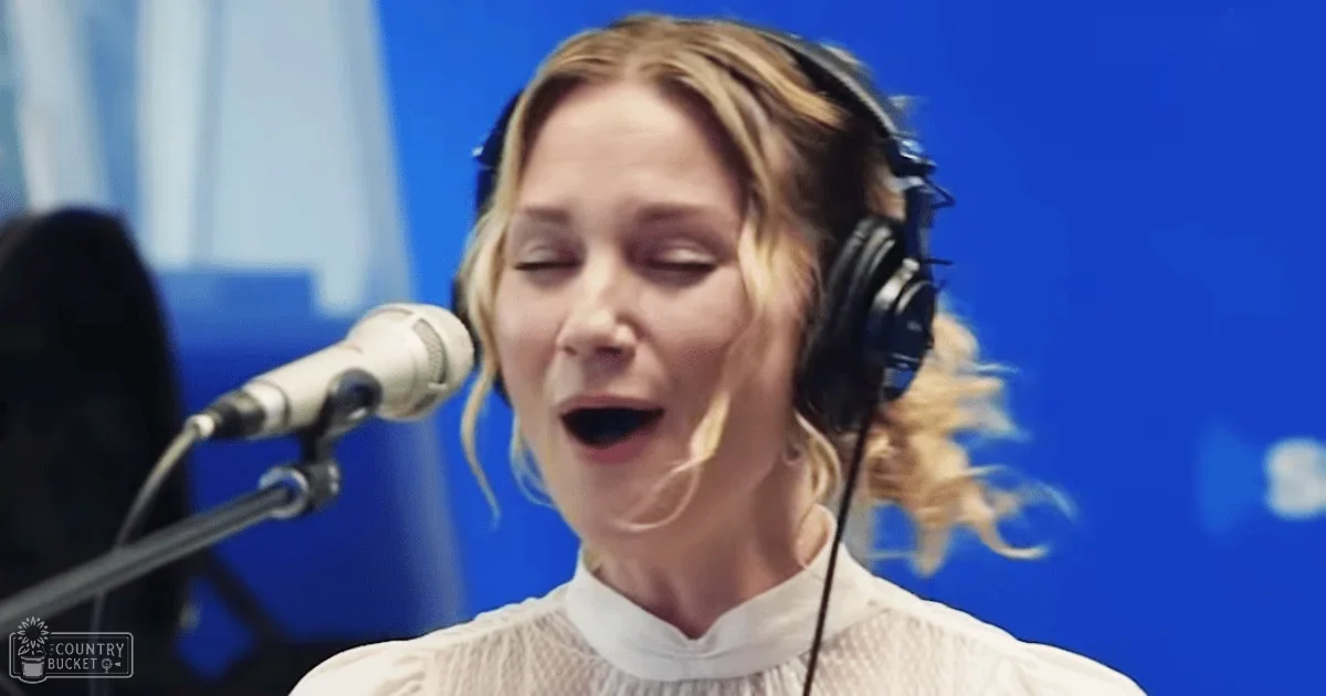 A young adult female wearing a white, ruffled-neckline blouse and large black over-ear headphones stands in front of a microphone in a professional recording studio with blue panels.