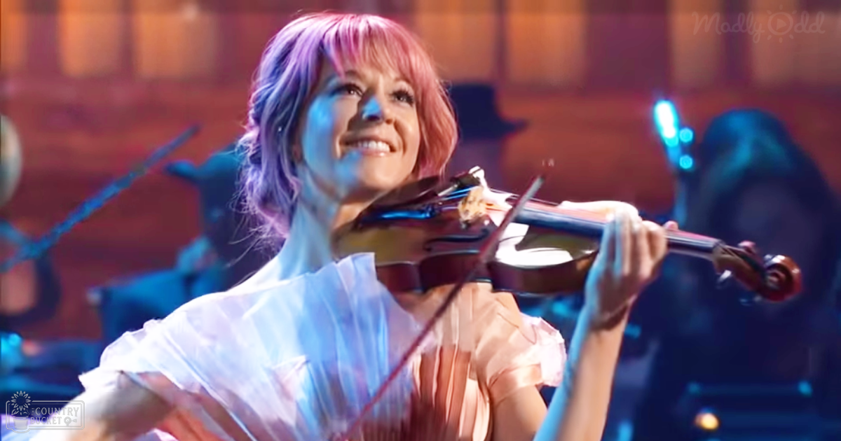 A young adult female wearing a light pink blouse with ruffled sleeves plays a classic wooden violin on stage during an evening concert under blue and warm lighting.