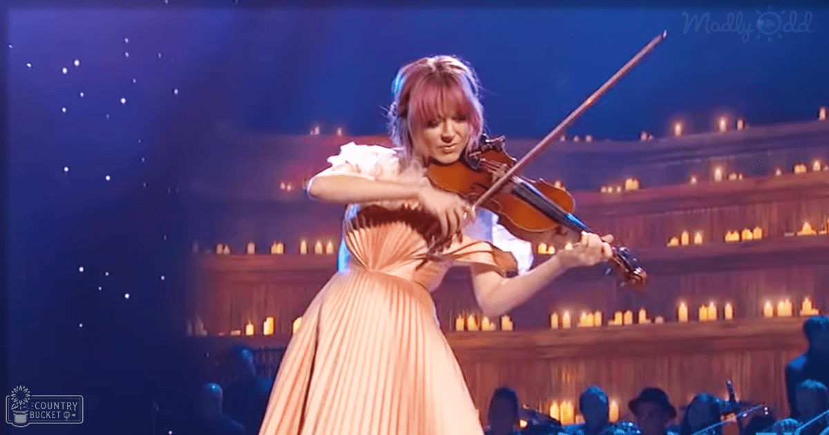 A young adult female violinist in an elegant, modern classical dress performs during a concert with candles and star-like lights in a dark blue backdrop.