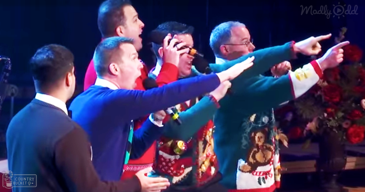 A group of adults in festive attire, possibly at a holiday performance or event, wearing suits and sweaters with holiday-themed designs. A bouquet of red flowers is visible.