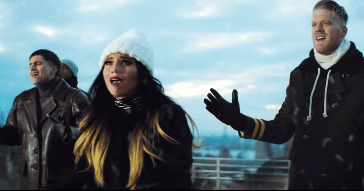 Young adult female singing with two male companions on an urban rooftop, partly cloudy sky suggesting late afternoon or early evening, winter setting due to heavy jackets and hats.
