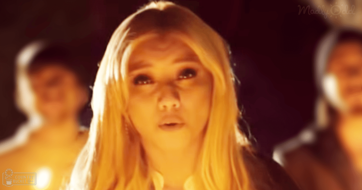 A young adult female with long blonde hair stands indoors wearing a white high-necked garment in a dimly lit environment with shades of yellow and orange lighting.