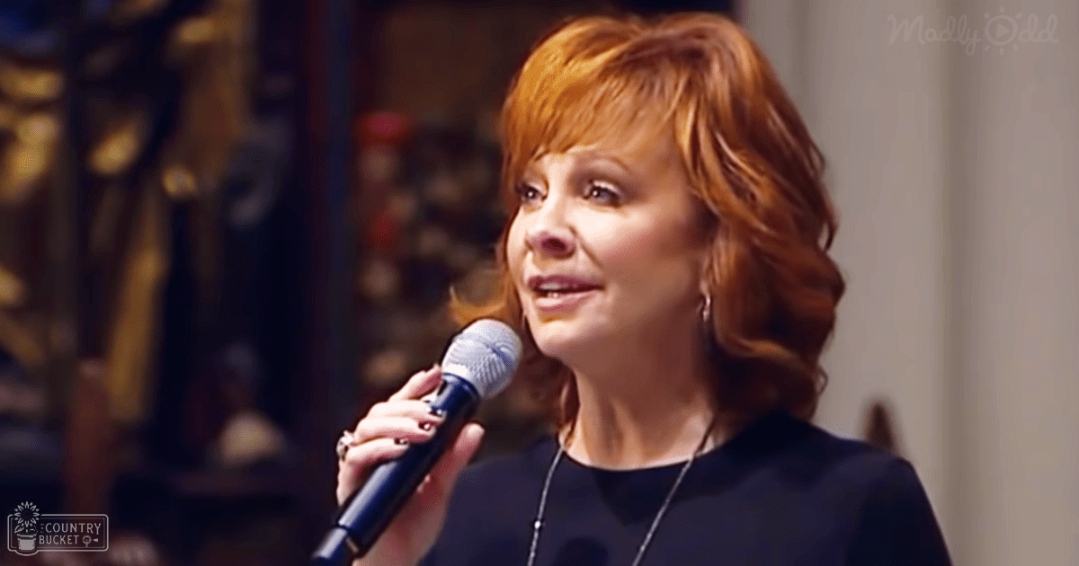 A middle-aged female singer wearing a dark-colored, short-sleeved, round-neck top with rings and a necklace, performing into a microphone indoors at night.