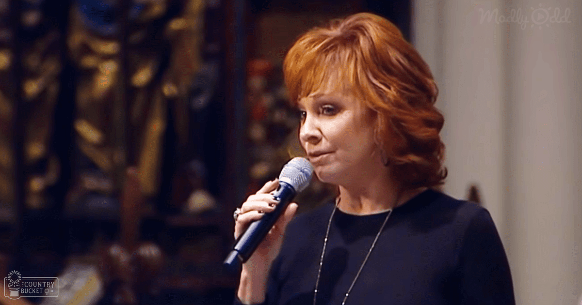 A middle-aged woman speaks into a microphone, wearing a dark-colored modern top indoors with warm lighting and an ornate background.