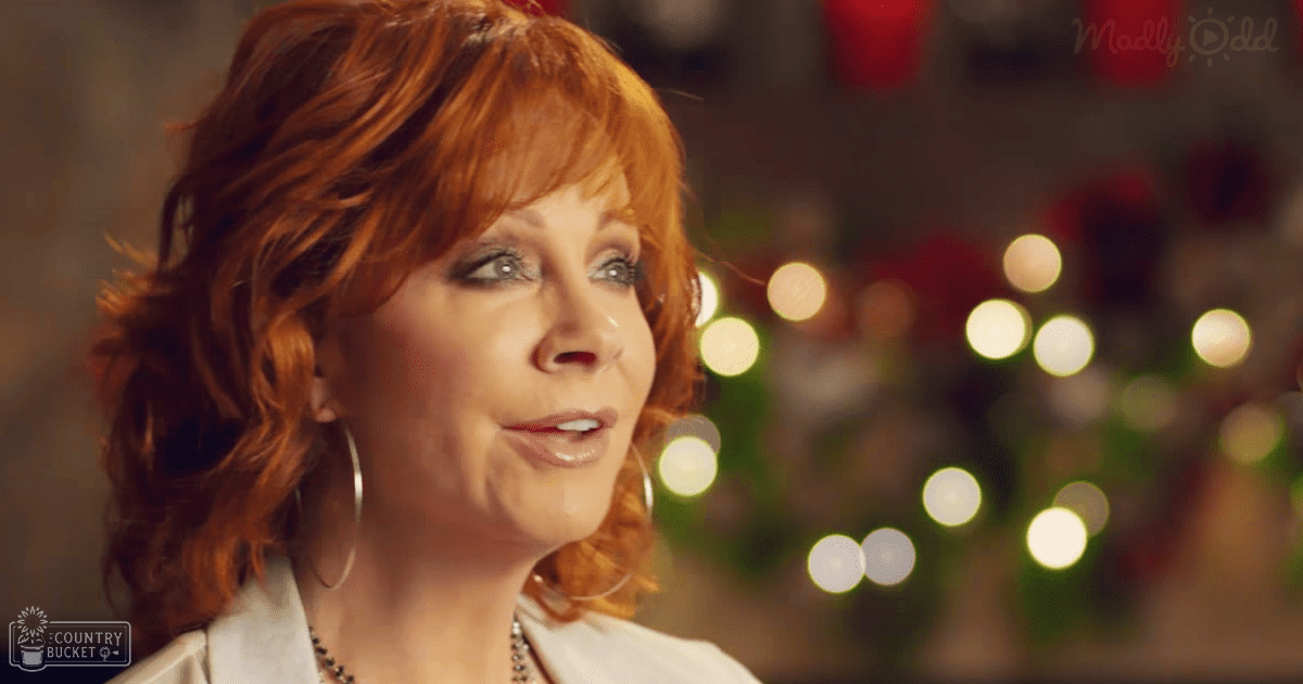A middle-aged adult female in a white, high-necked blouse with short sleeves, wearing large hoop earrings and a necklace with small beads, standing indoors adorned with festive Christmas decorations.