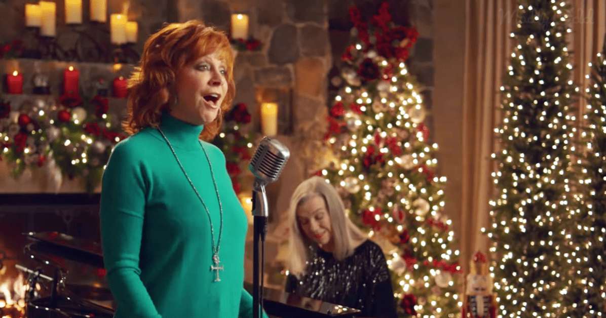 A middle-aged woman singing in front of a Christmas tree with a vintage microphone and piano keys visible, indoors during the holiday season.