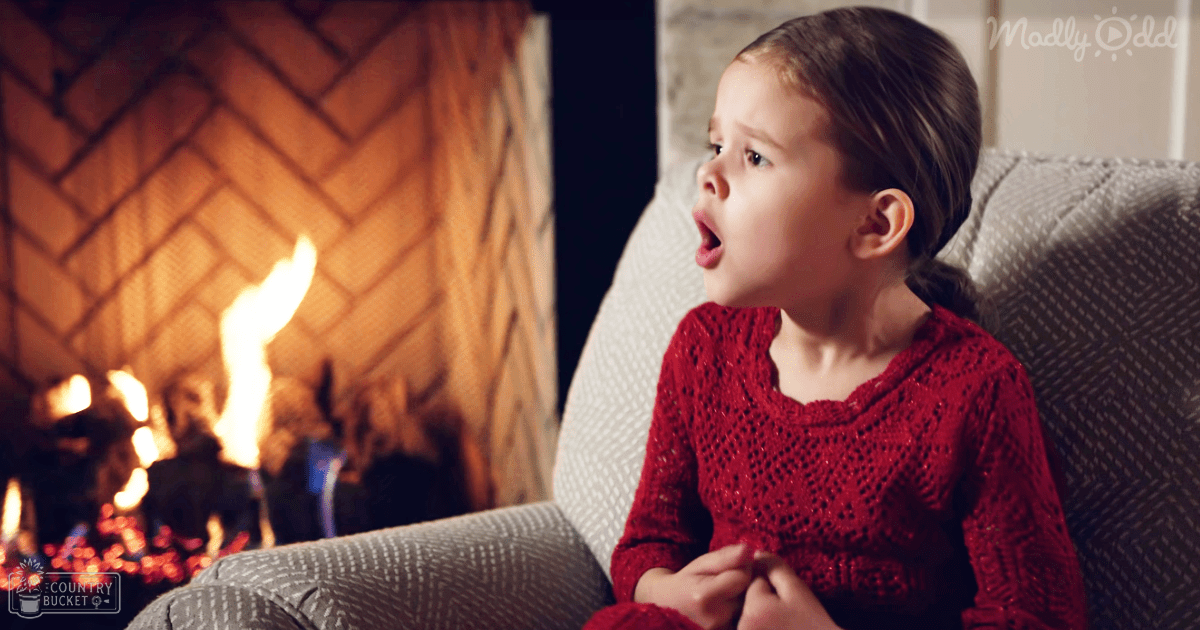 A toddler girl wearing a red knitted sweater stands indoors by a lit brick fireplace, with a gray upholstered chair and warm lighting, suggesting an evening winter setting.