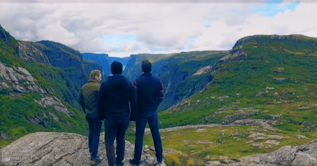 Three individuals stand at a viewpoint overlooking a mountainous landscape in Norway, wearing casual outdoor attire.