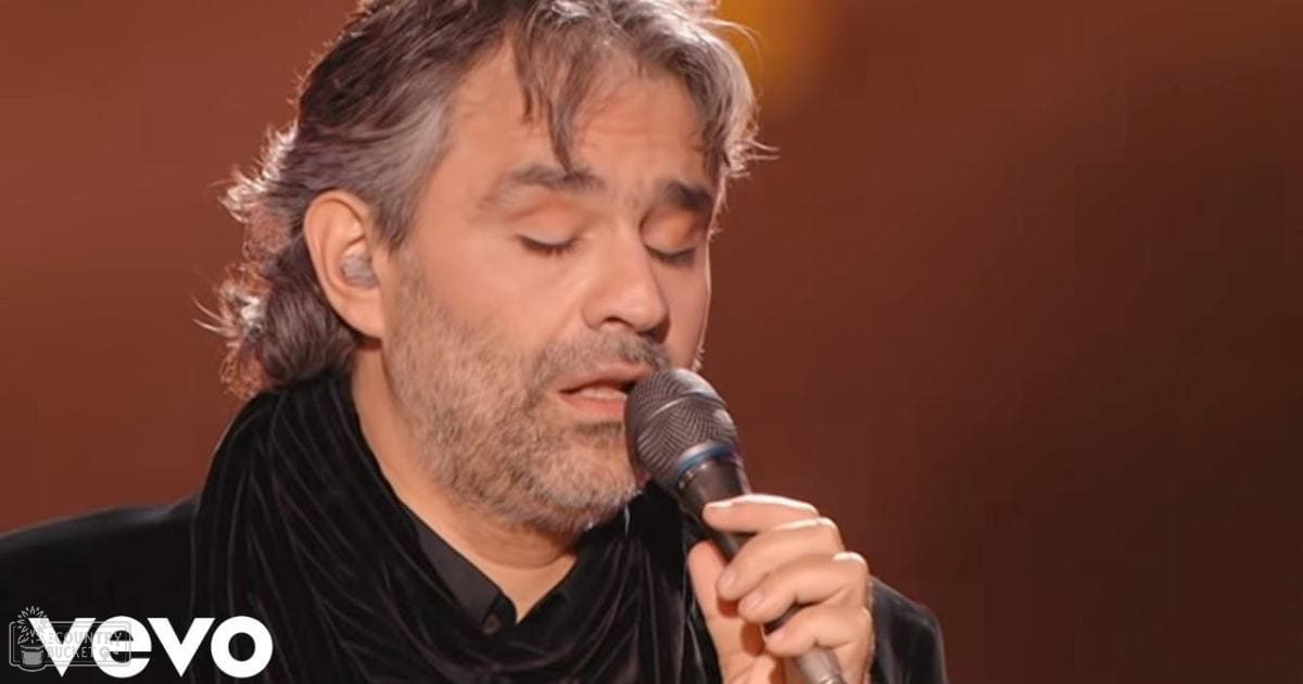 Middle-aged adult male singer wearing a black high-collared garment with a draped scarf around his neck, holding a handheld microphone on an indoor stage backdrop.