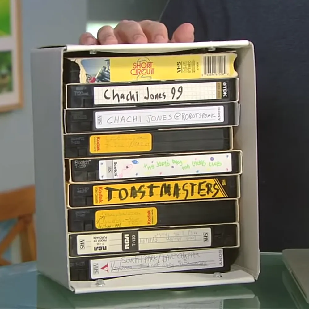 Stack of VHS tapes with handwritten titles in a white cardboard box, indoor setting, natural light, daytime