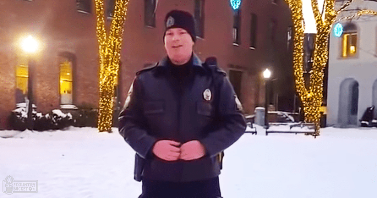 A young adult male in a dark blue police uniform stands outdoors at night with festive lighting, snow on the ground, and illuminated brick buildings in the background.