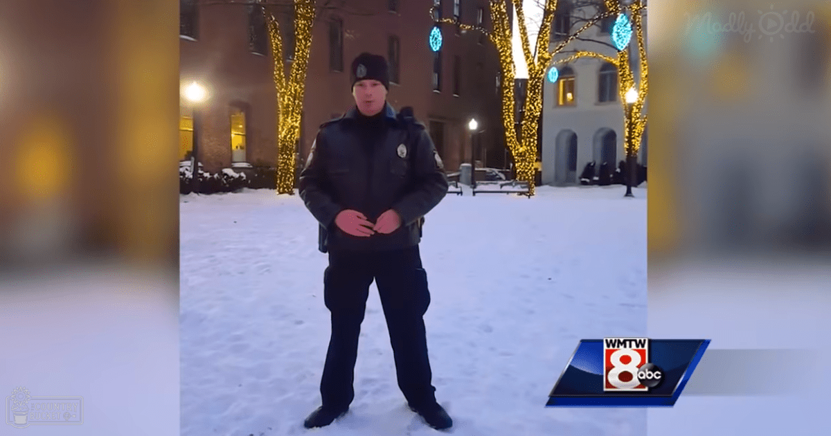 An adult male police officer wearing a dark uniform jacket with patches and black dress shoes stands outdoors on a snowy evening in front of a building decorated with lights, possibly indicating an institutional or university setting.