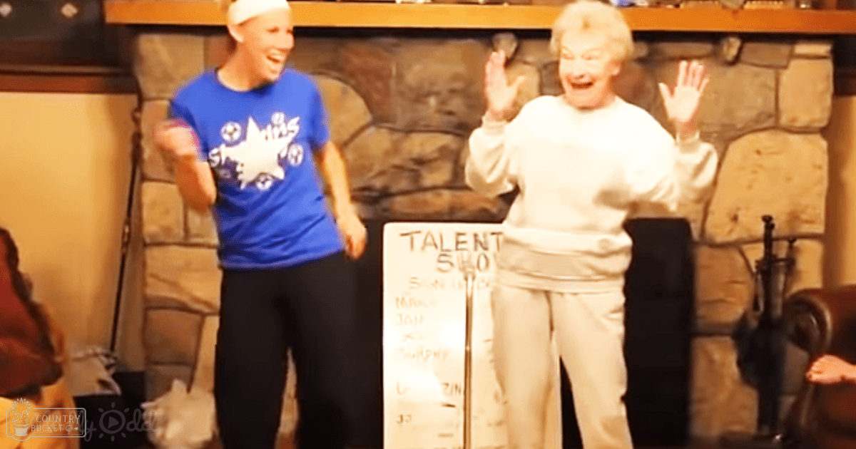 A talent show event indoors with young adult female in a blue t-shirt and older adult female in white top, set against stone walls and a cozy fireplace ambiance.