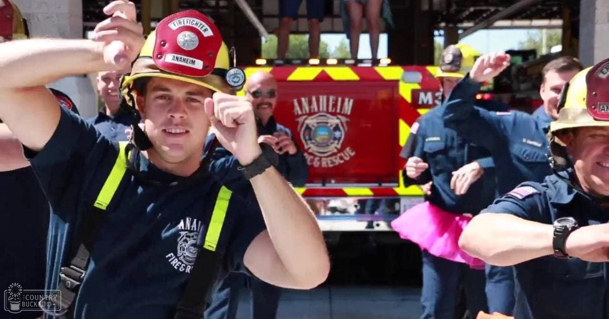 A young adult male firefighter wearing a navy blue 'Anaheim Fire & Rescue' shirt, yellow reflective safety vest, red helmet with 'Firefighter Anaheim', amidst other firefighters at an elevated platform during what appears to be a public event in Anaheim, California.